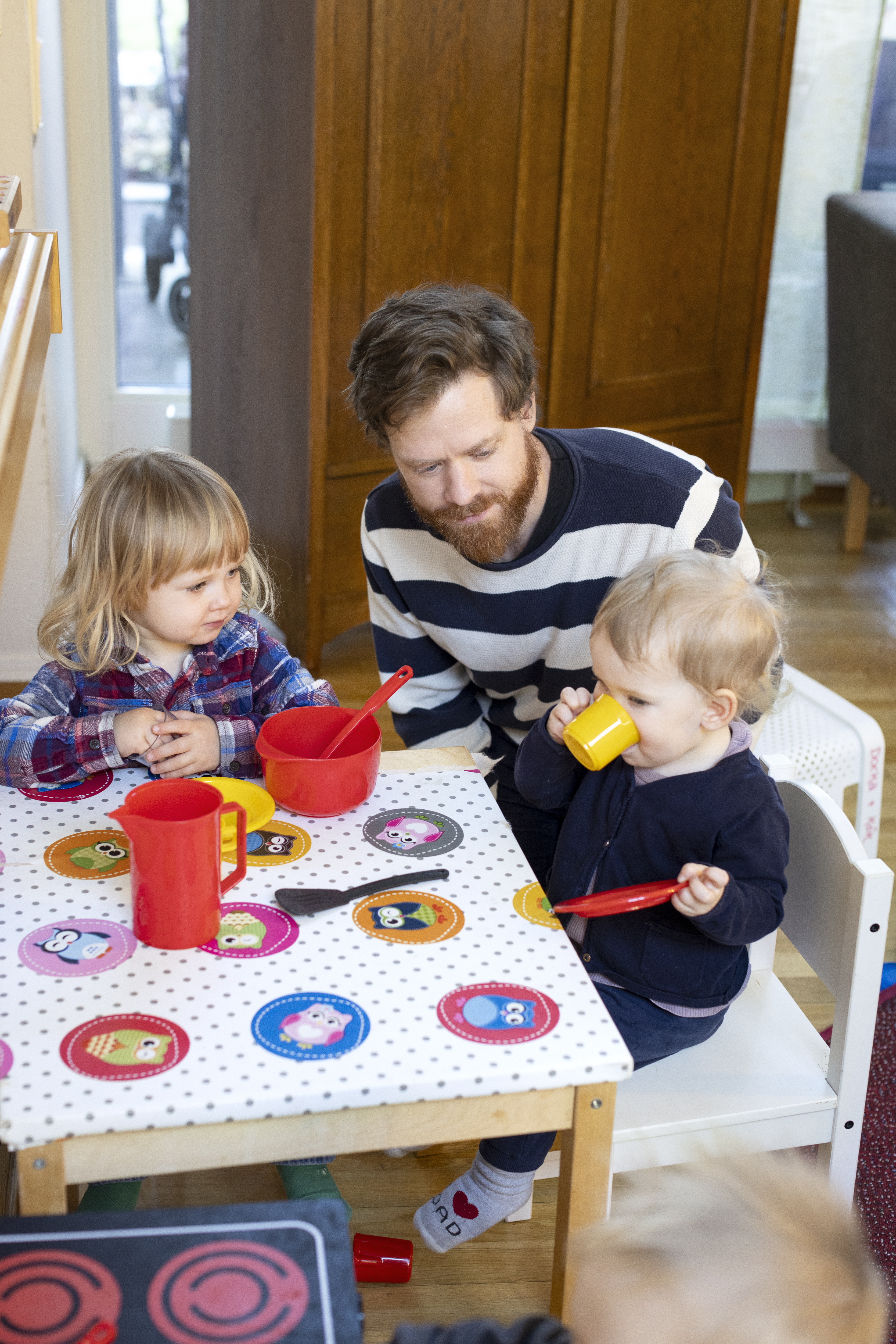 En man sitter vid ett barnbord tillsammans med två små barn. Ett av barnen låtsas-dricker ur en liten gul plastmugg.