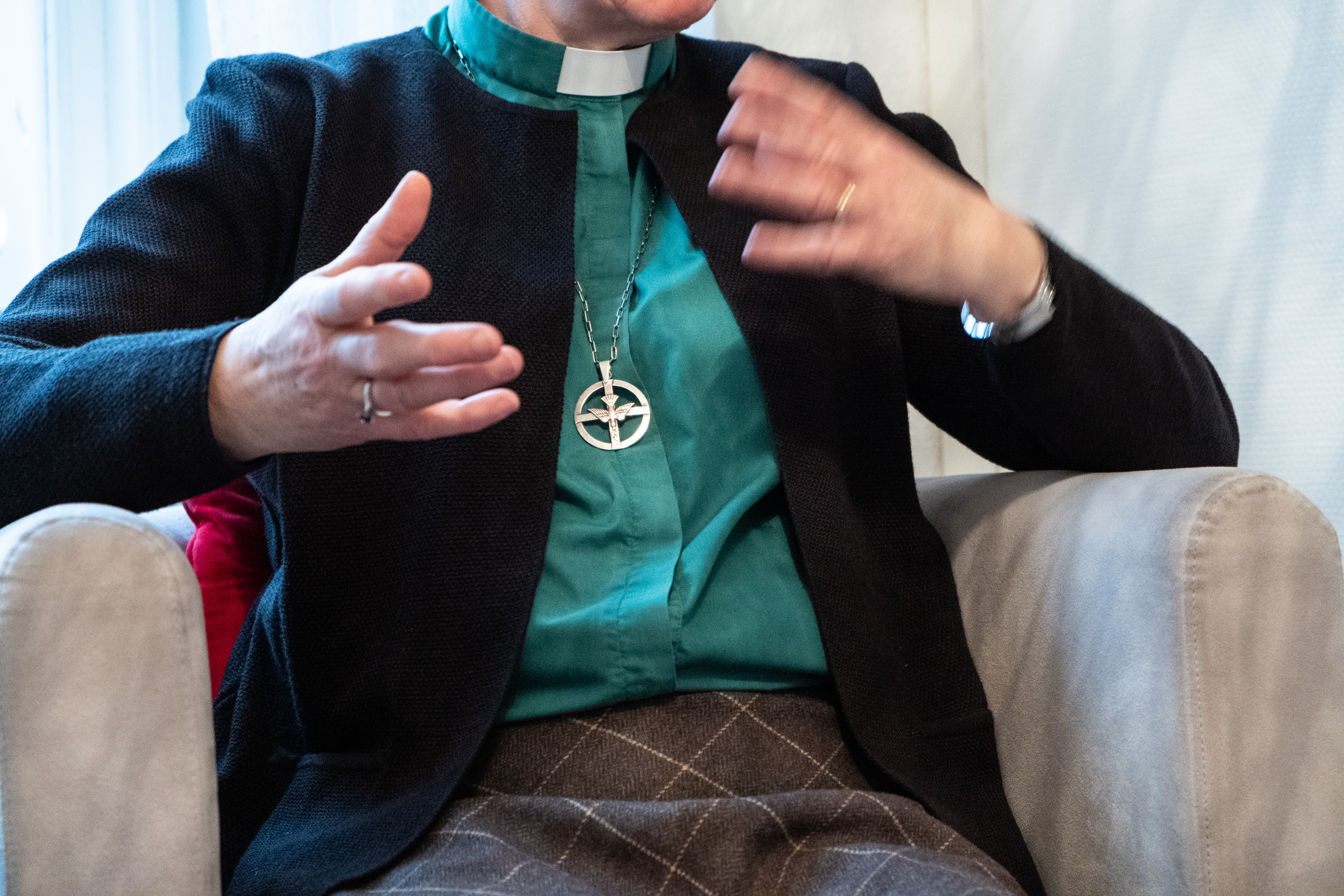 En diakon sitter i en fåtölj och gestikulerar. Hen bär diakonemblemet runt halsen.