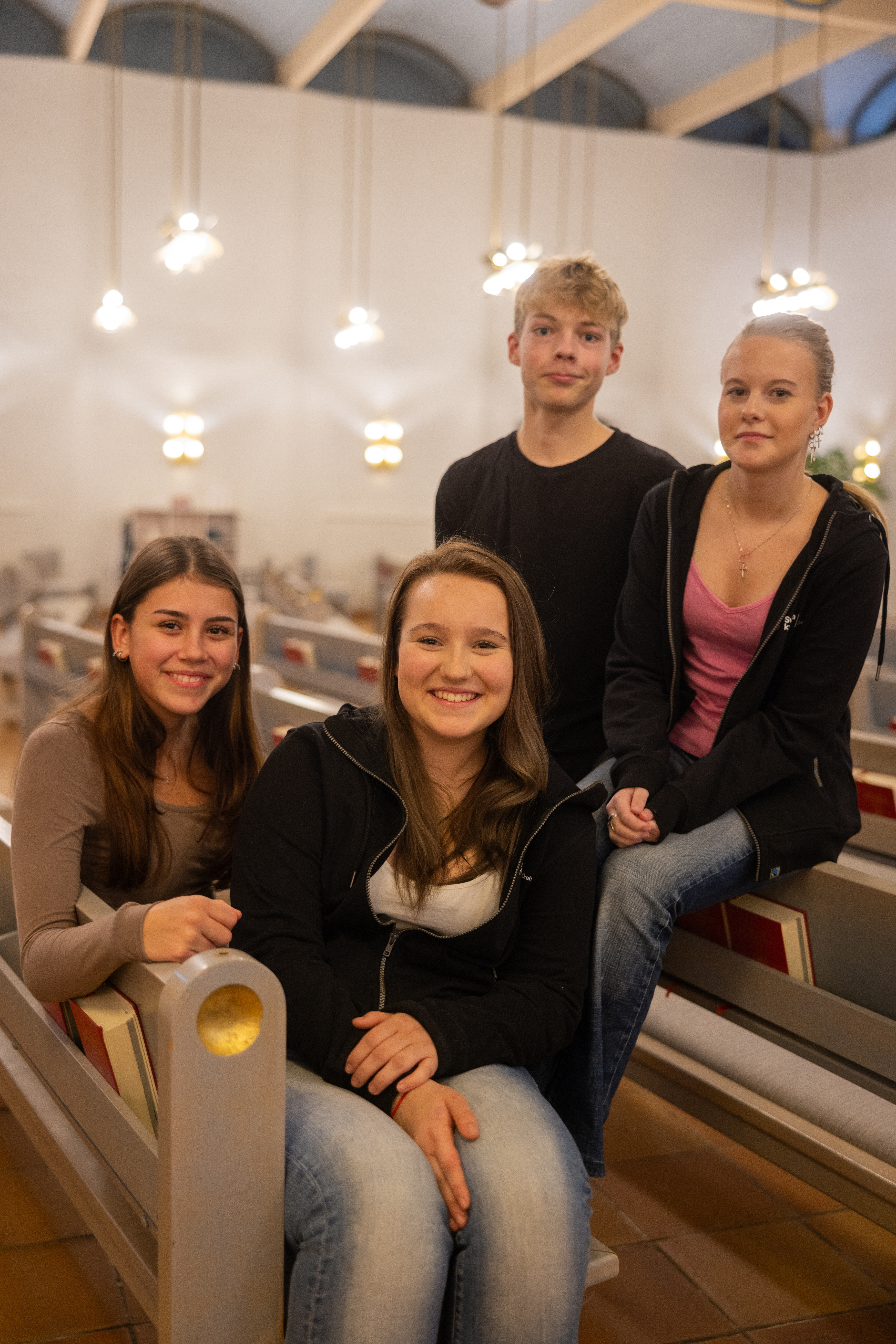 Fyra glada ungdomar sitter i kyrkbänken och tittar in i kameran.