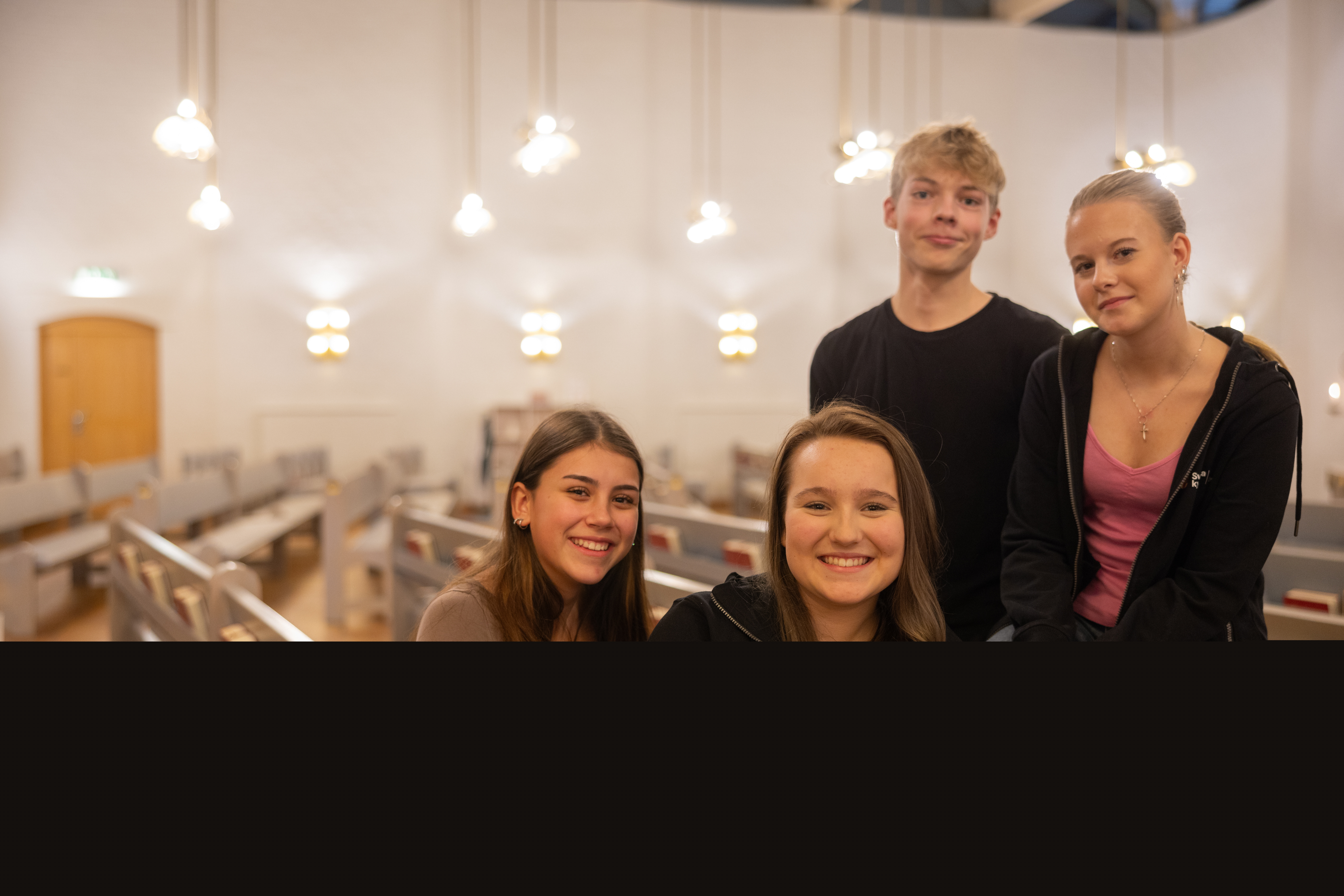 Fyra glada ungdomar sitter i kyrkbänken och tittar in i kameran.