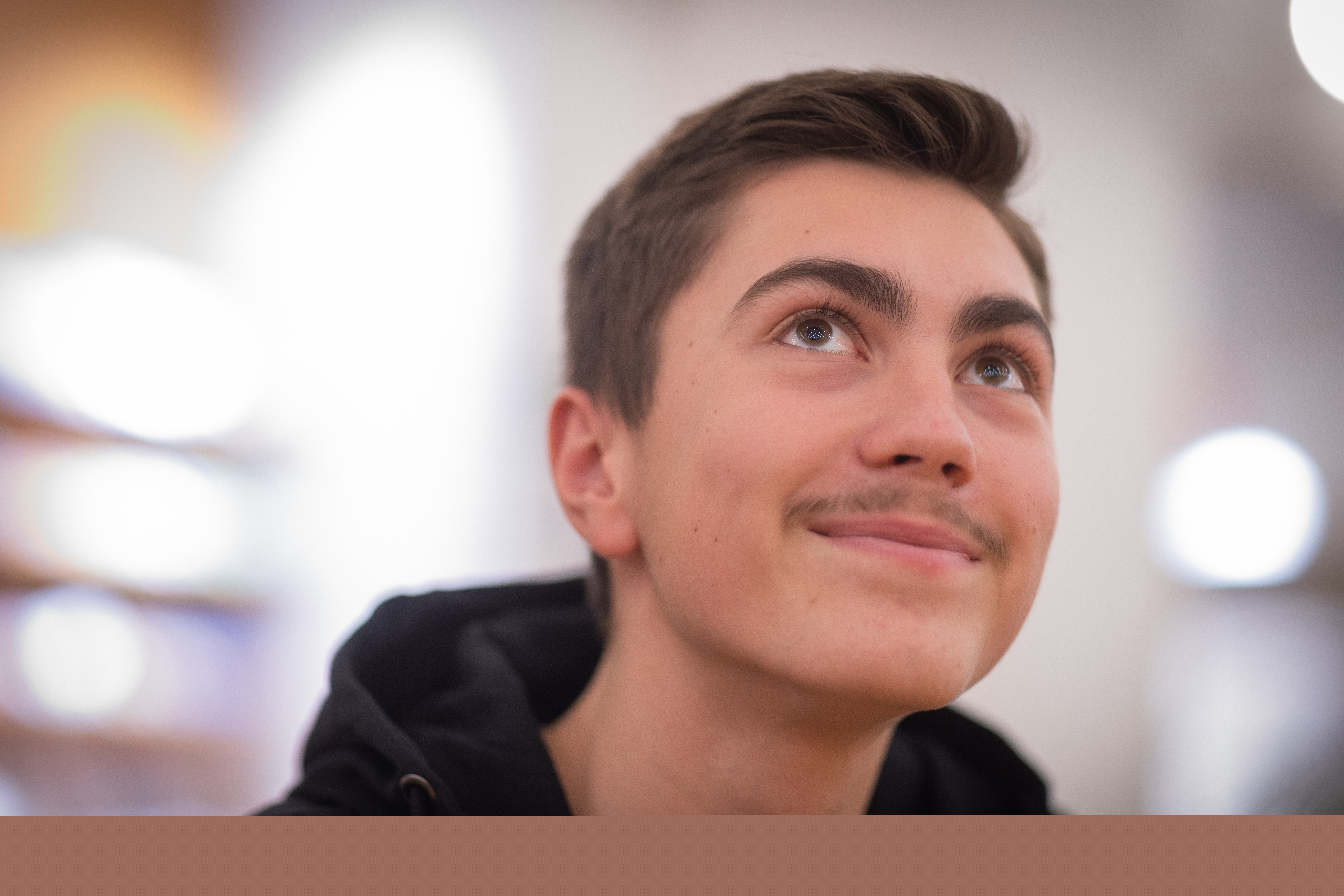 Närbild på en ung leende kille i svart luvtröja som blickar uppåt. Solljus strimmar in från fönstret bakom honom.