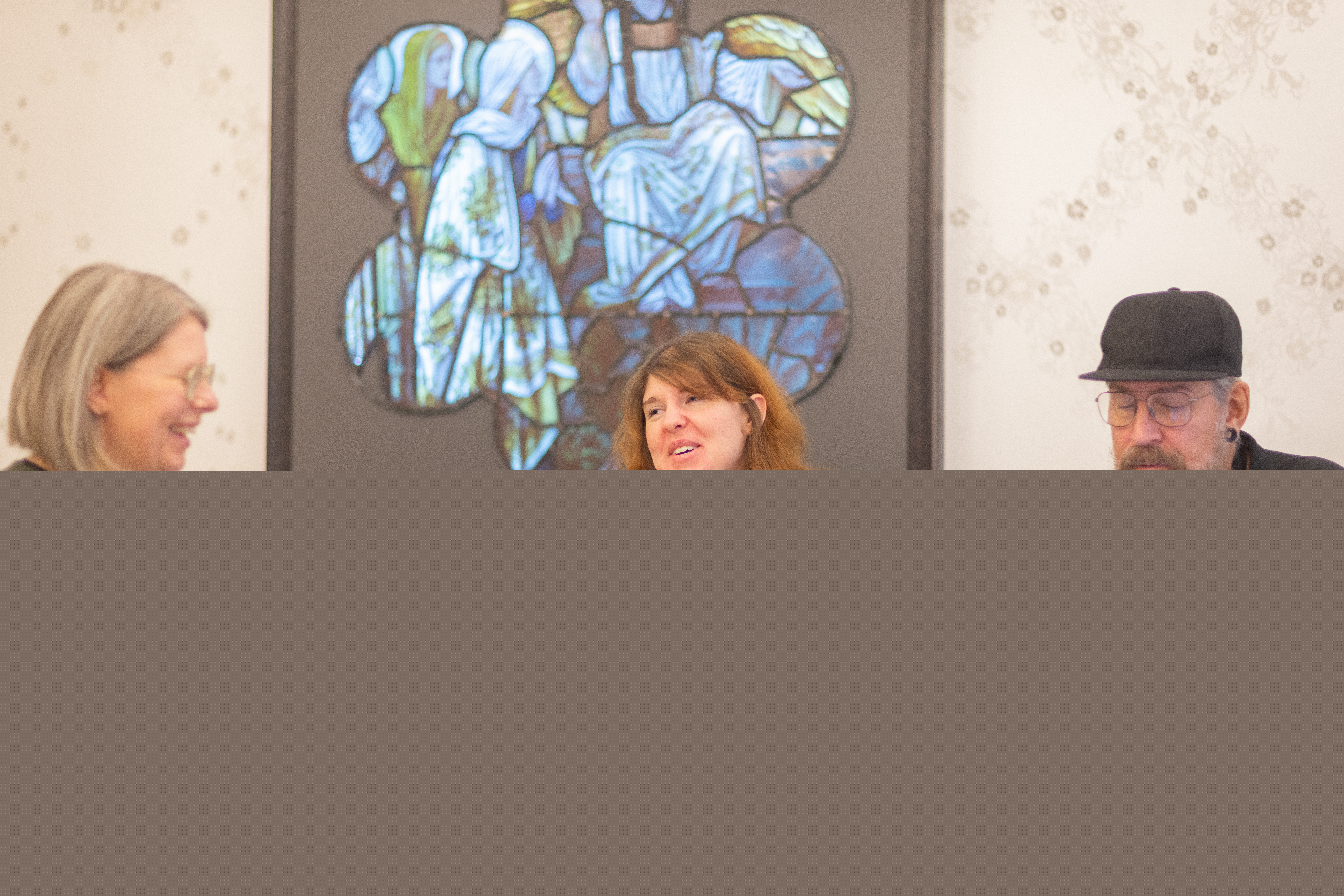 Två kvinnor och en man sitter och samtalar kring ett bord. På väggen bakom hänger en tavla i blå och gröna färger.