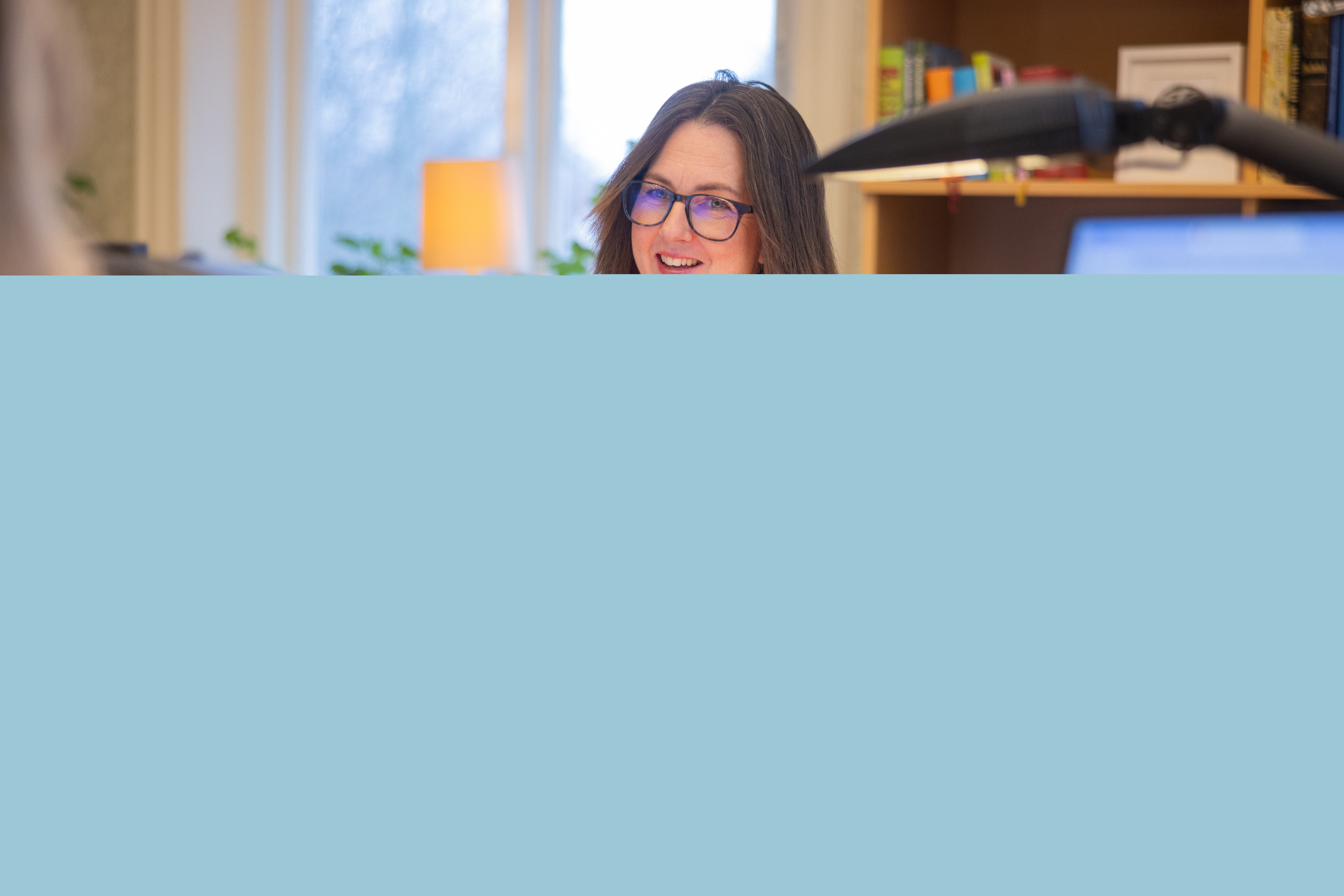 En kvinna i beige tröja sitter och arbetar på ett kontor. Hon pratar med en kollega.