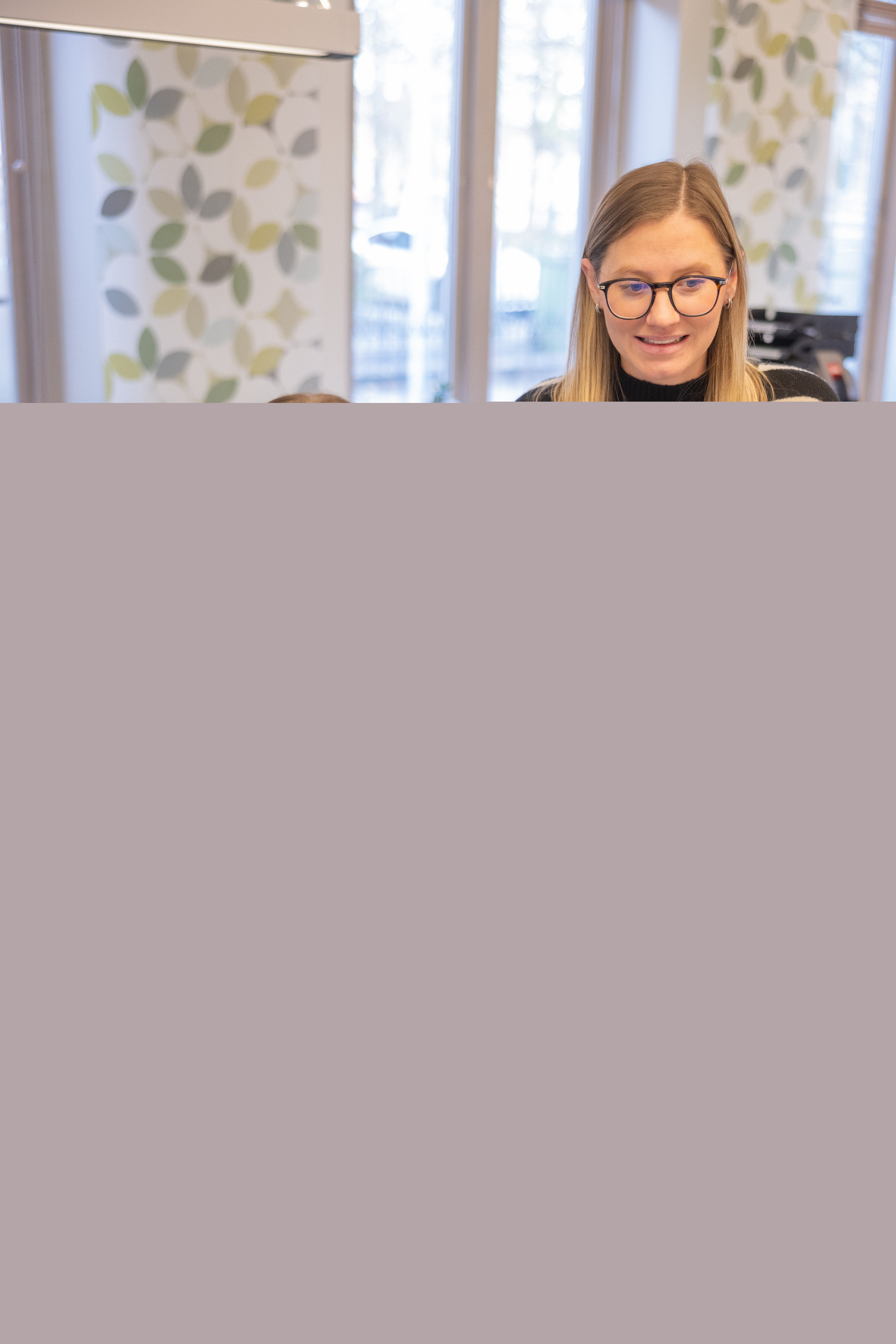 Två kvinnor framför en dator i en reception. Vita gardiner med gröna löv i bakgrunden.