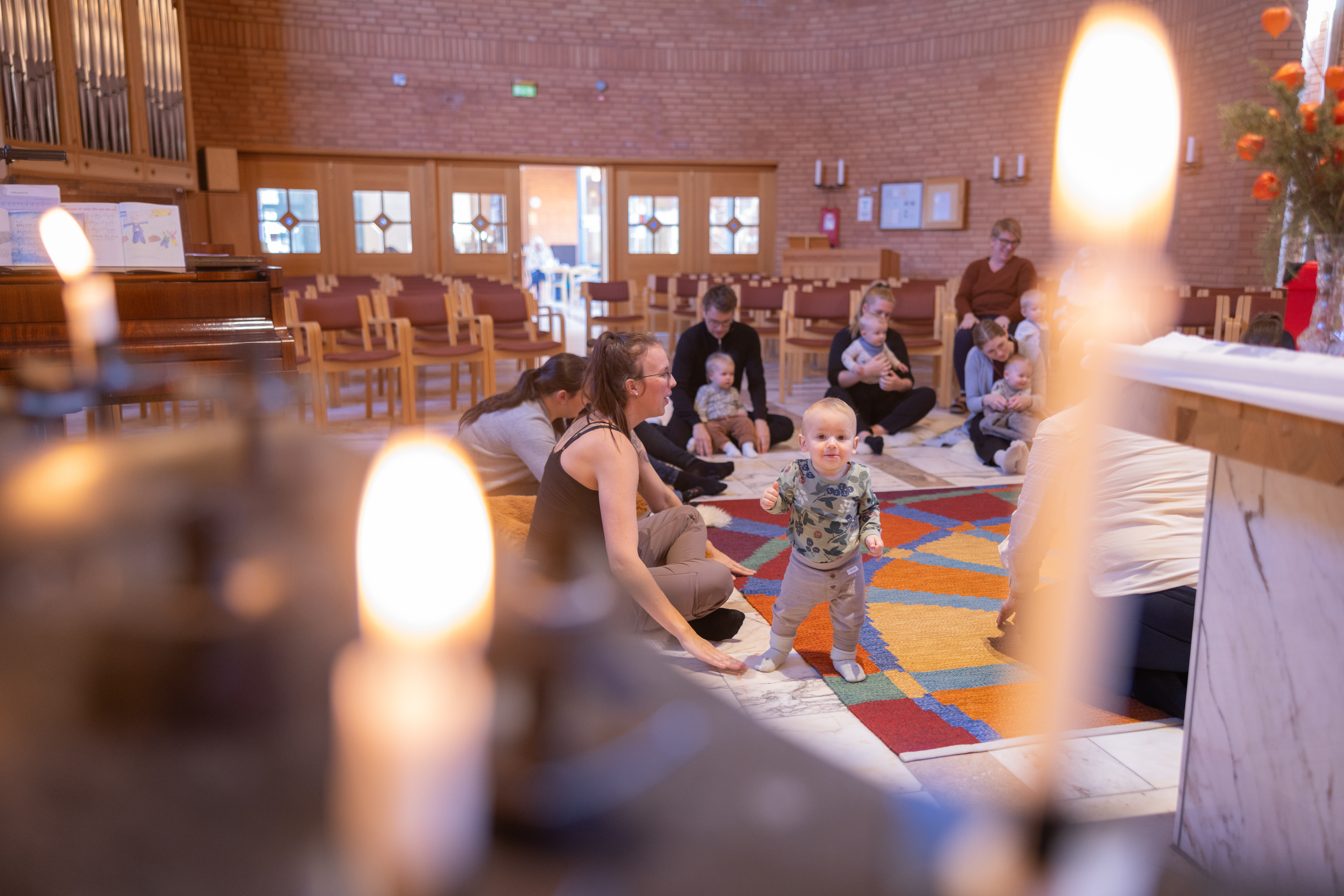Vuxna och barn sitter på golvet i en kyrka och har sångstund på en öppen förskola. Två tända ljus i förgrunden.