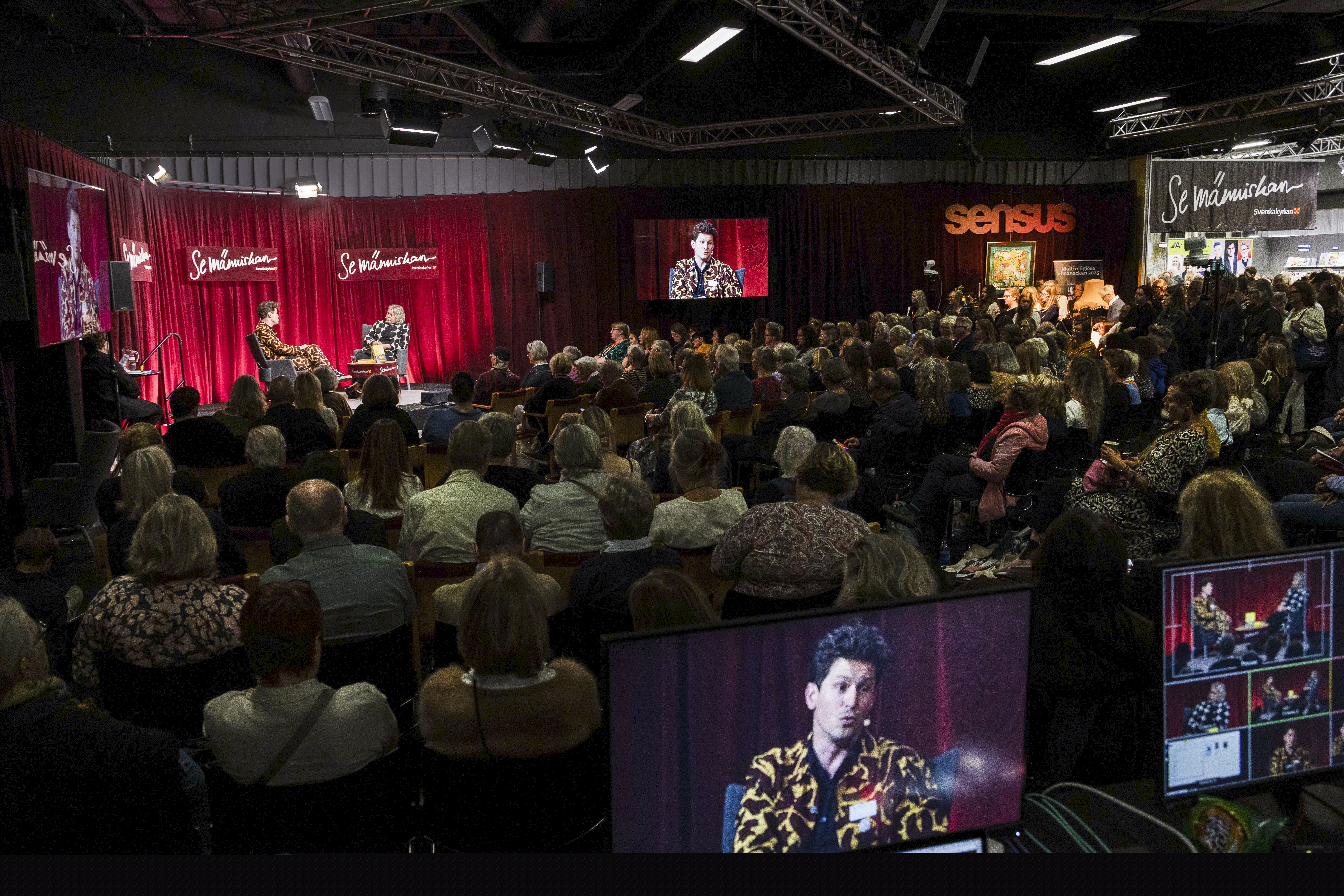 Sittande publik i en lokal på bokmässan. På scenen pågår ett samtal mellan två personer.