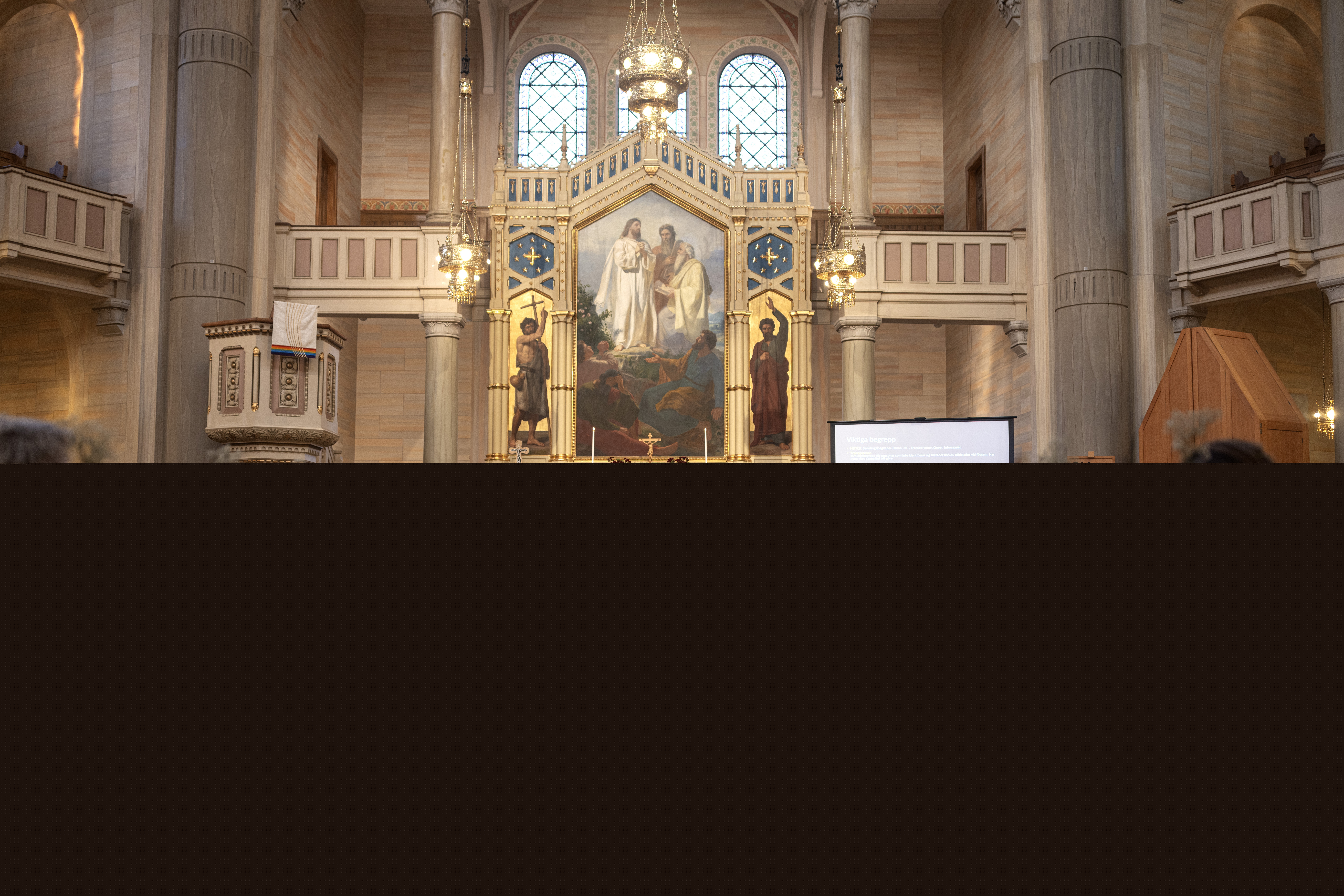 En präst och en yngre kvinna står vid ett bord längst fram i en kyrka och håller ett föredrag. Åhörare sitter i kyrkbänkarna. En regnbågsfärgad matta i mittgången.