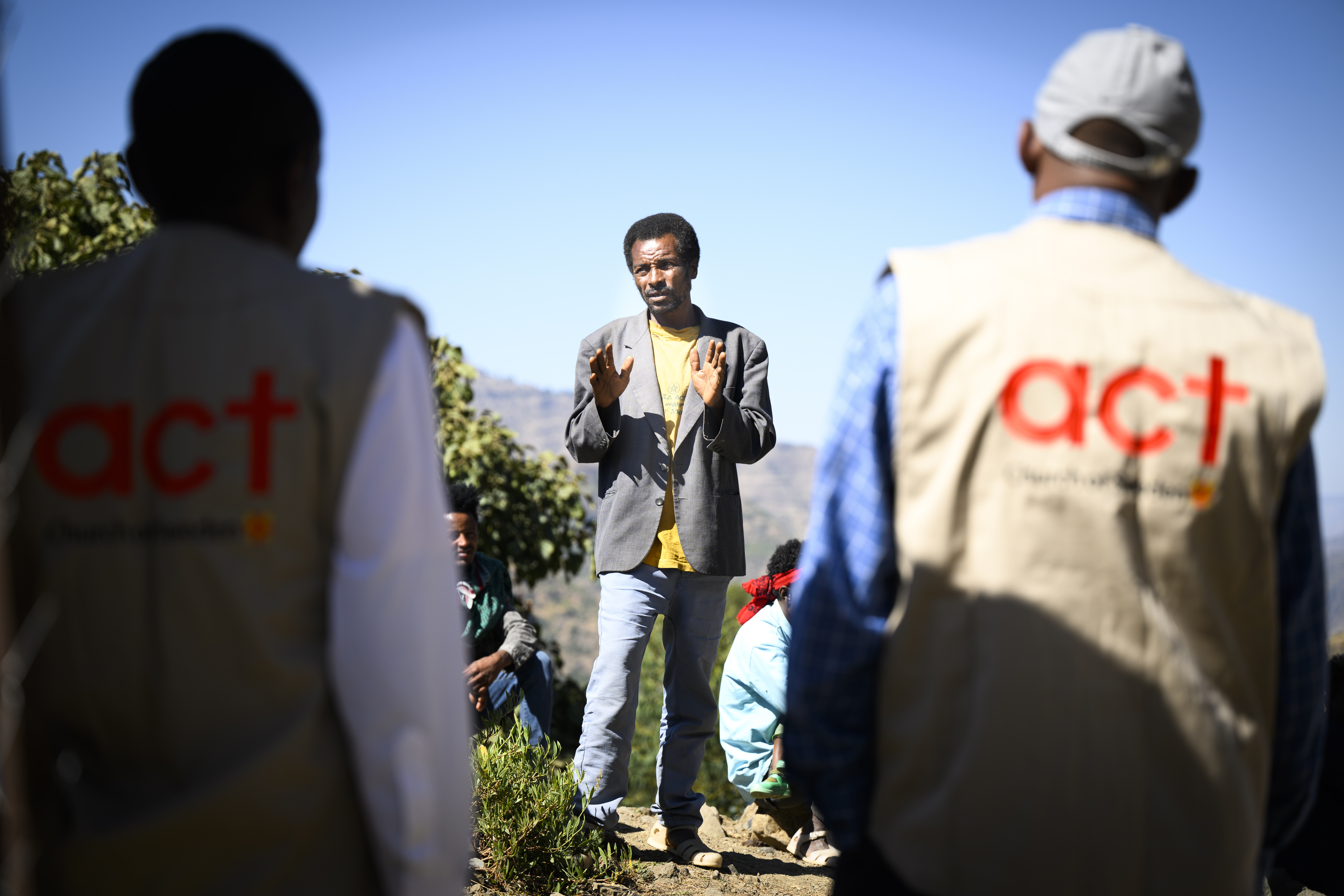 En man står och förklarar något, i förgrunden syns två personer med Act Svenska kyrkans logotyp på ryggen. 