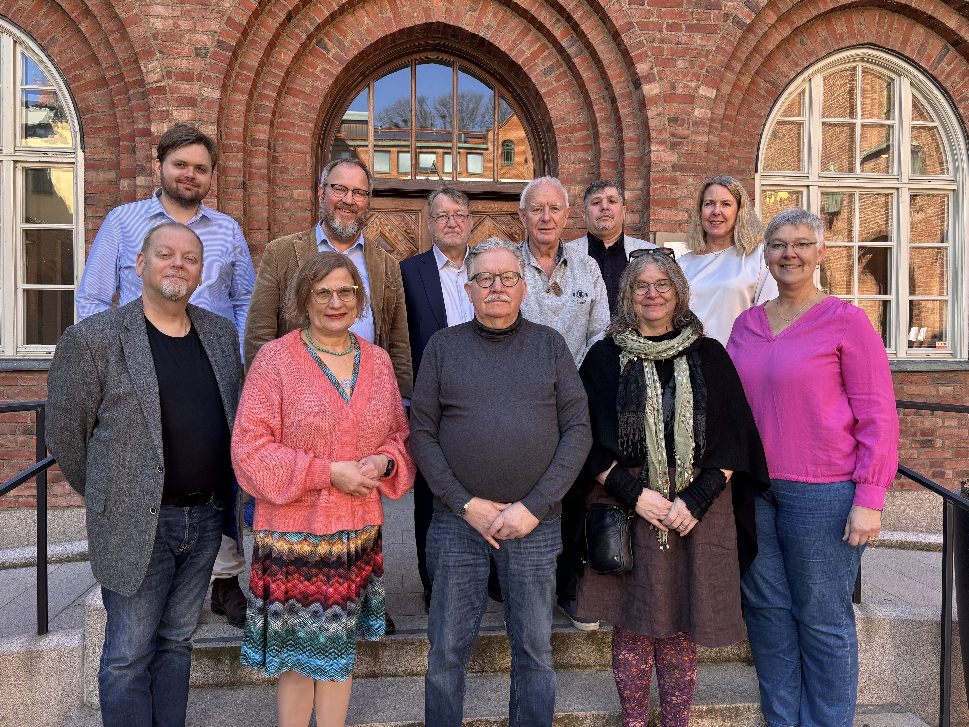 Gruppfoto med elva vuxna personer, både män och kvinnor, som står tätt tillsammans på stentrappan framför en äldre byggnad i rött tegel. Bakom gruppen syns en rundad träport och höga fönster med vita karmar. Byggnaden har dekorativa tegelbågar som ramar in dörren och fönstren. Personerna är klädda i vardagskläder i olika färger och mönster, bland annat rosa, blått, grått och jordtoner. Några står på ett högre trappsteg bakom de andra, vilket skapar två rader. Det är dagsljus och miljön ger ett intryck av en historisk eller institutionell plats.