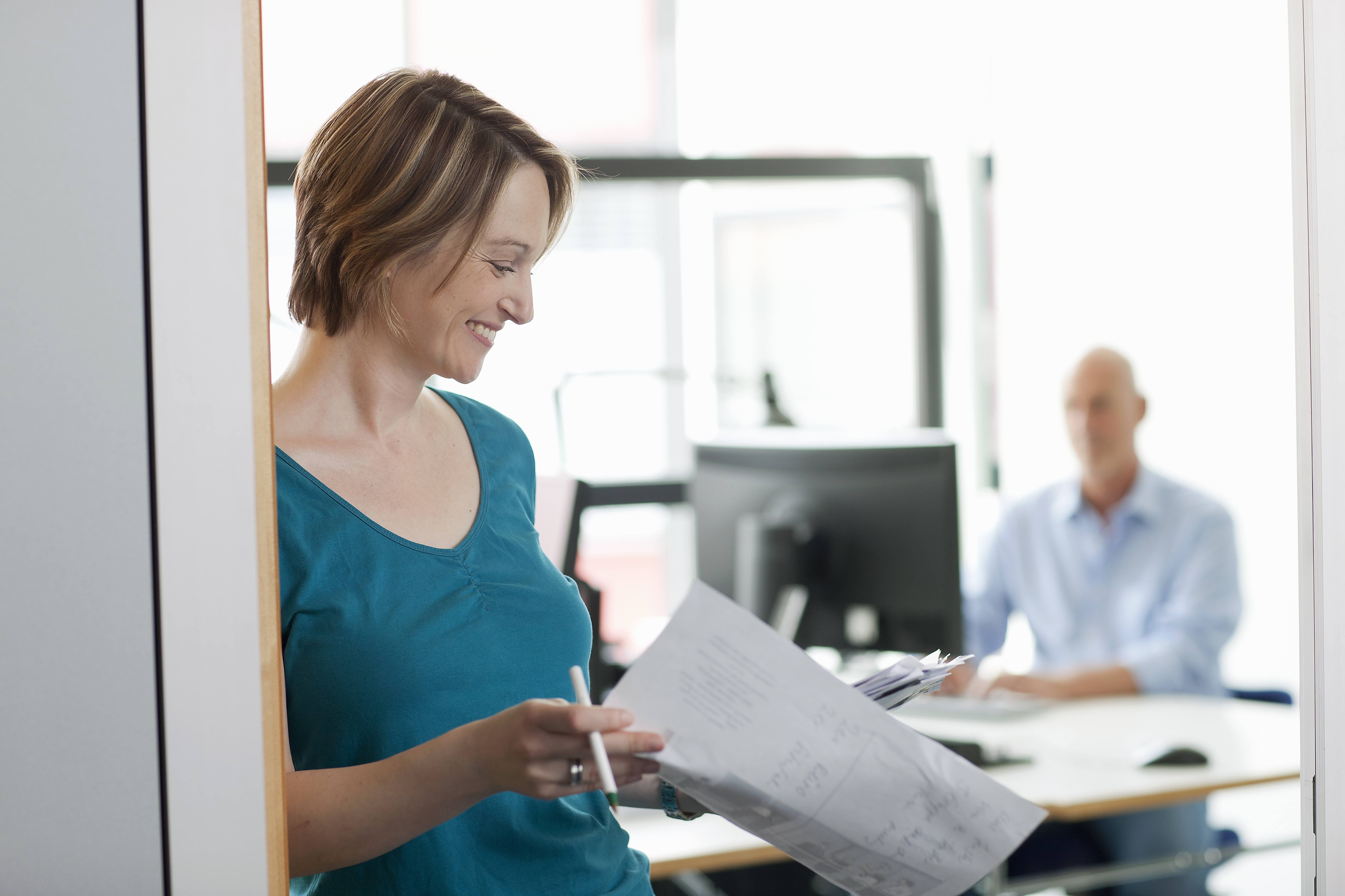 En kvinna med kort hår lutar sig mot en vägg och läser nöjt på ett papper. I bakgrunden syns suddigt en gestalt som arbetar vid en dator.
