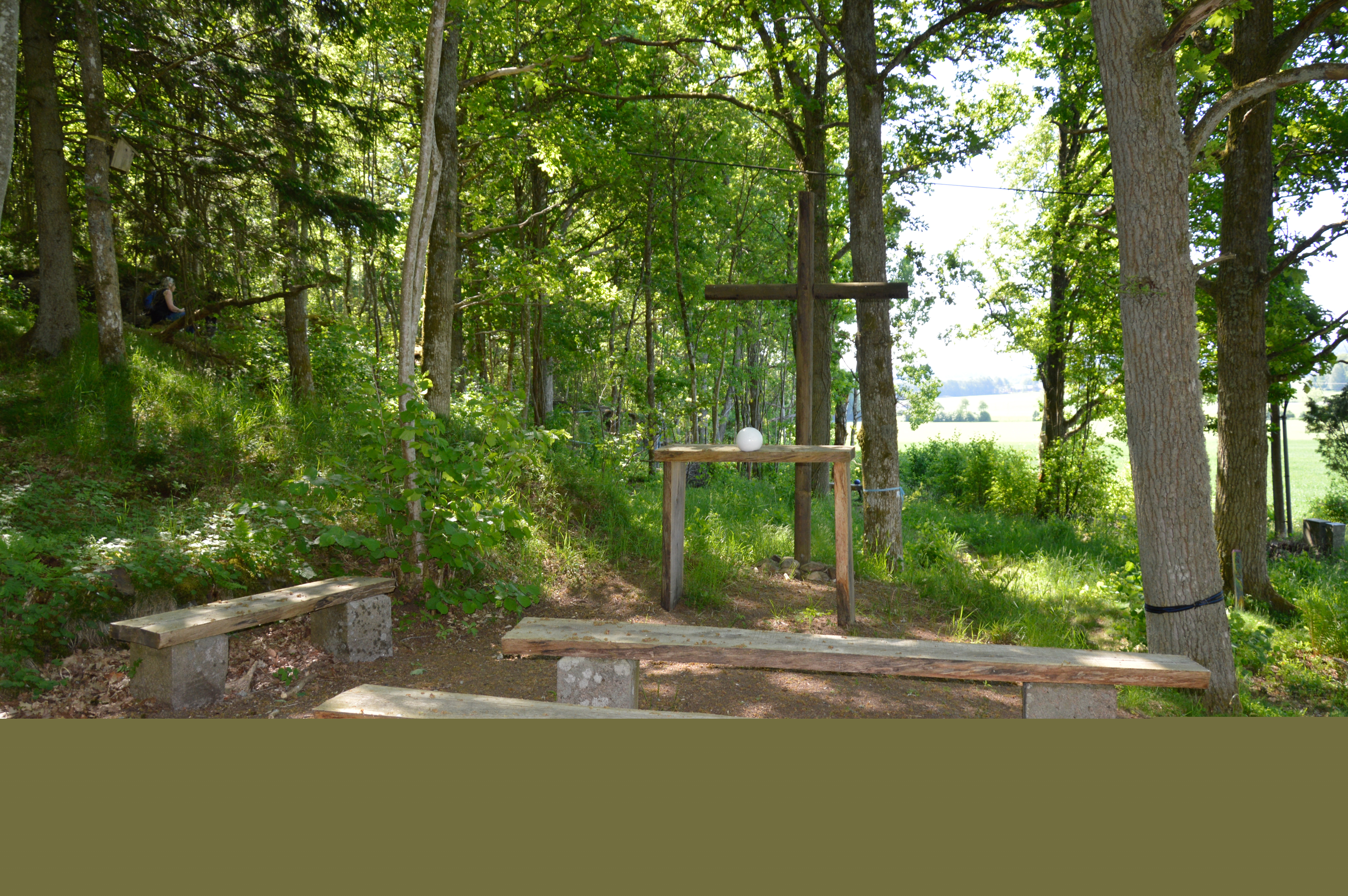 Altare, kors och kyrkbänkar utomhus i en skogsdunge. På altaret ligger en vit stor pärla från frälsarkransvandringen i Östad.