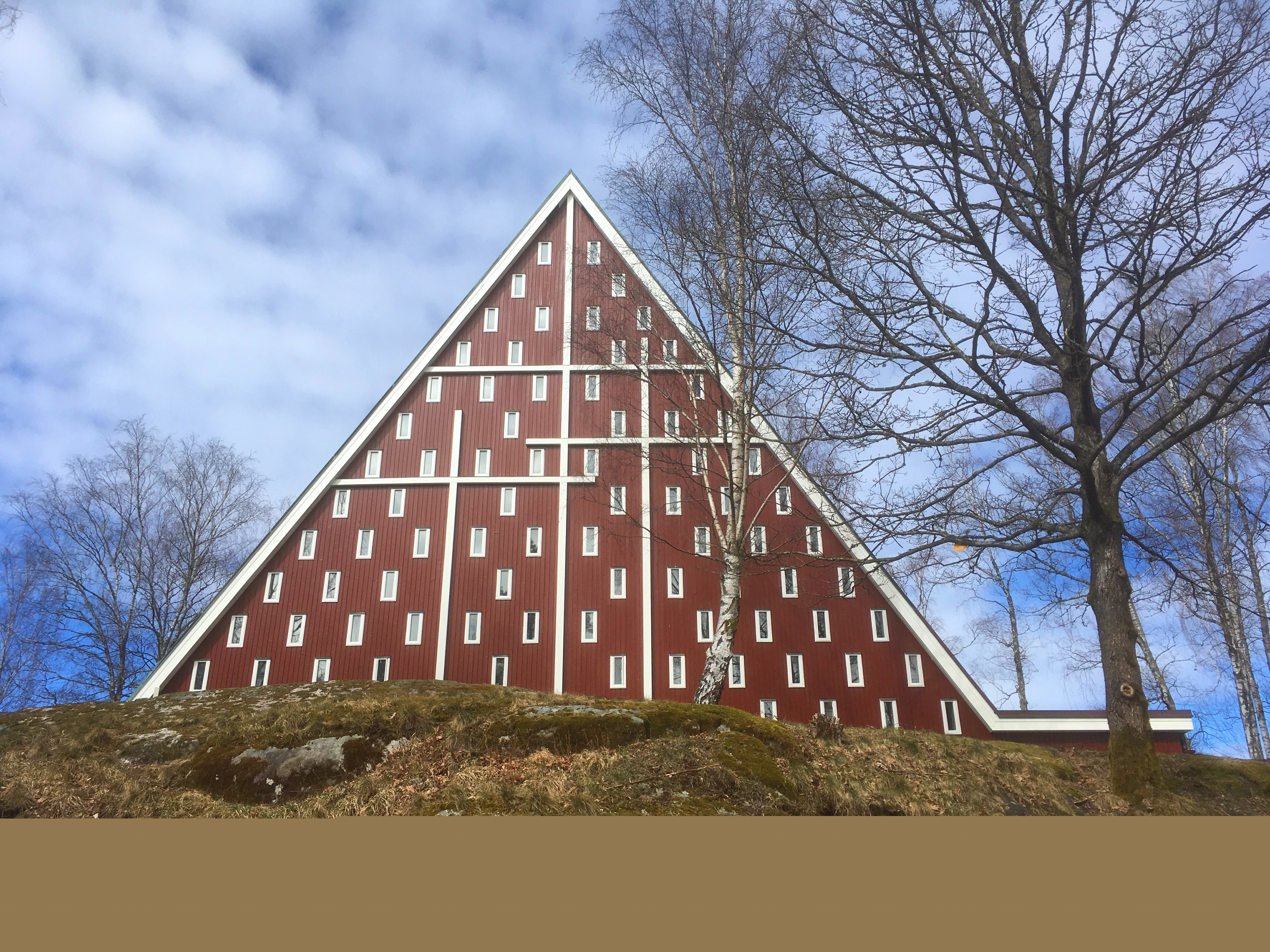 Röd träkyrka med tre vita kors på gaveln. Omgiven av träd utan löv.