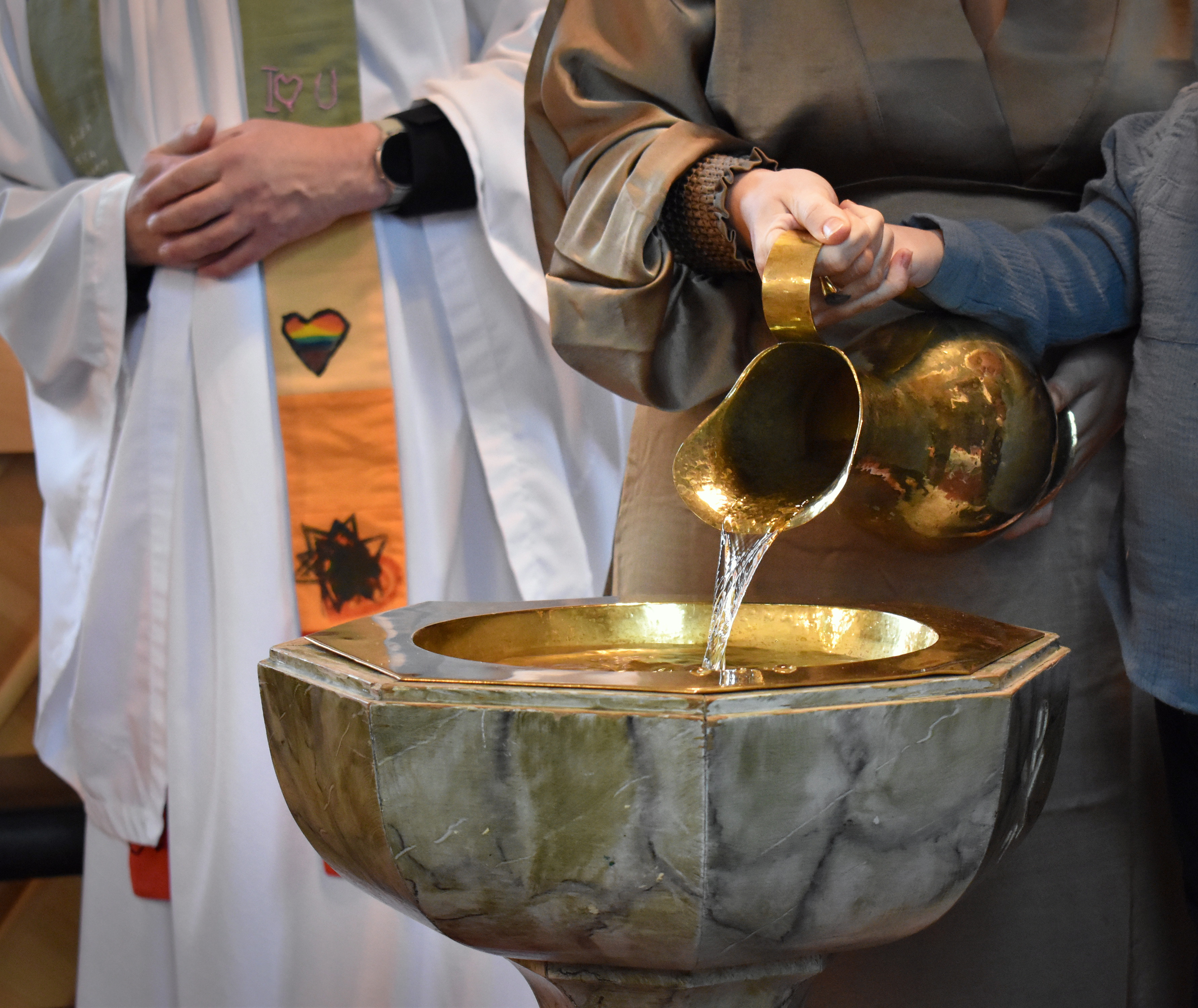 Pojke fyller dopfunt med vatten ur en kanna. Han får hjälp av en vuxen.