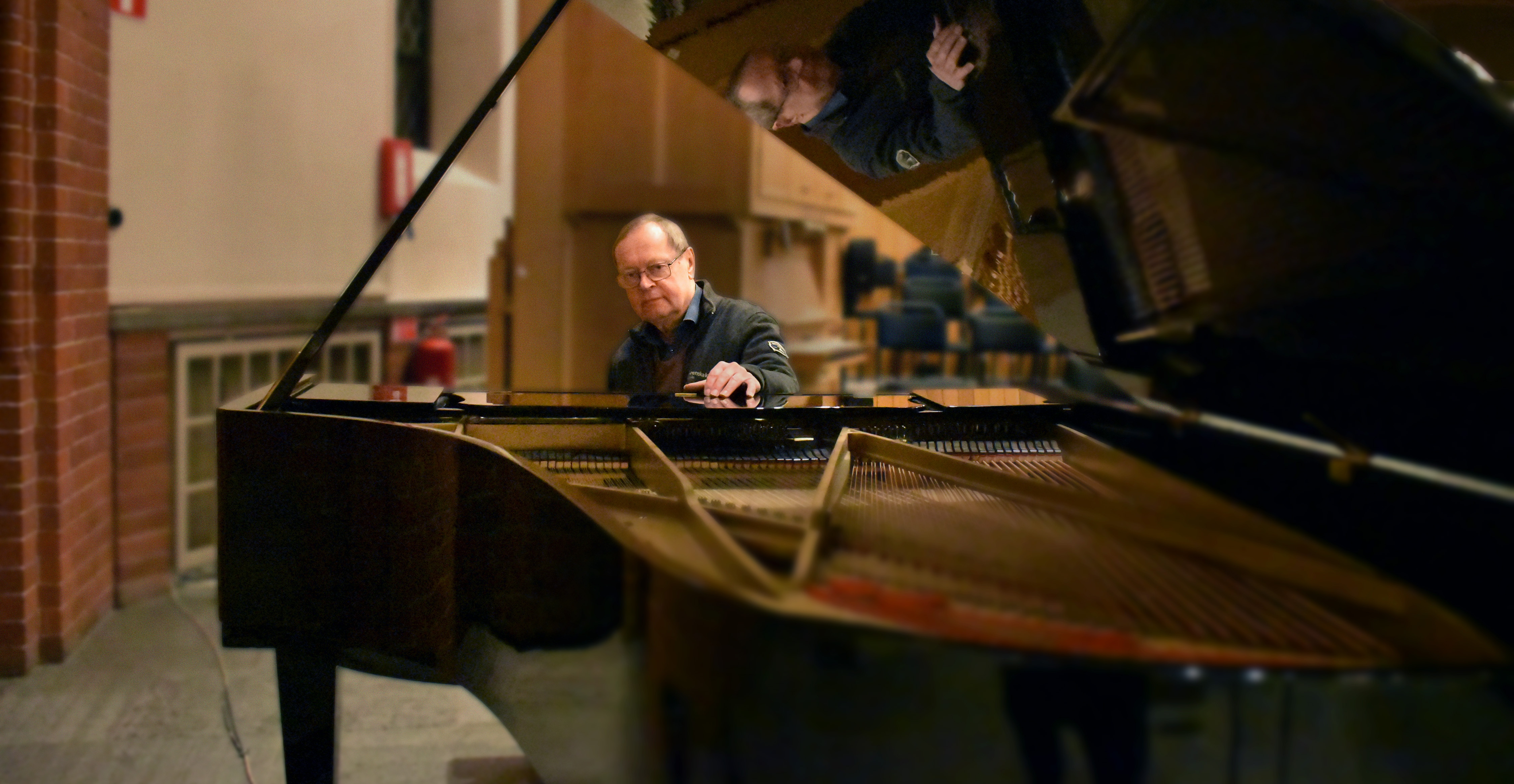Man med glasögon sitter vid flygel i kyrka.