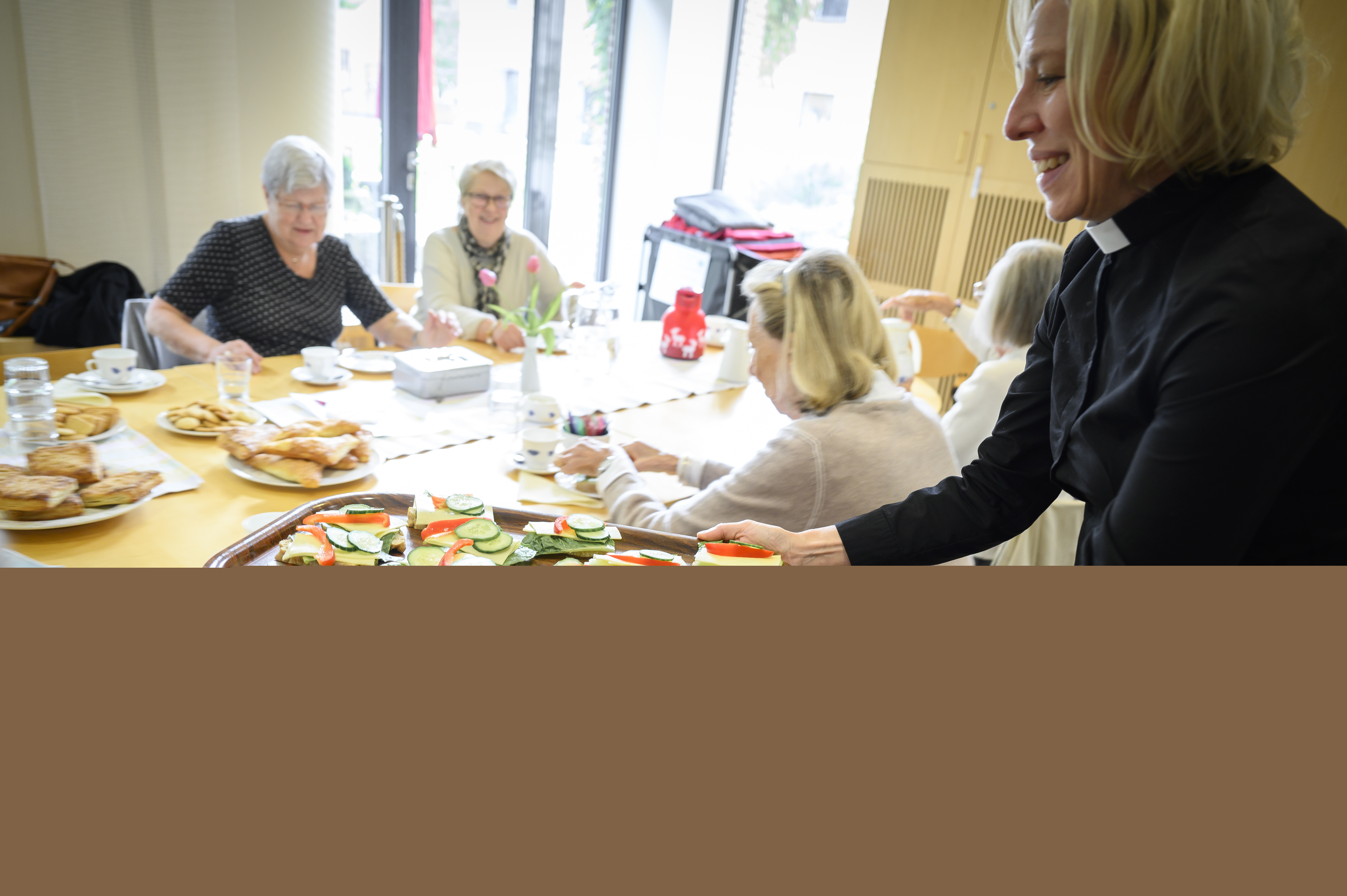 En kvinnlig präst delar ut smörgåsar till några damer som sitter runt ett bord.