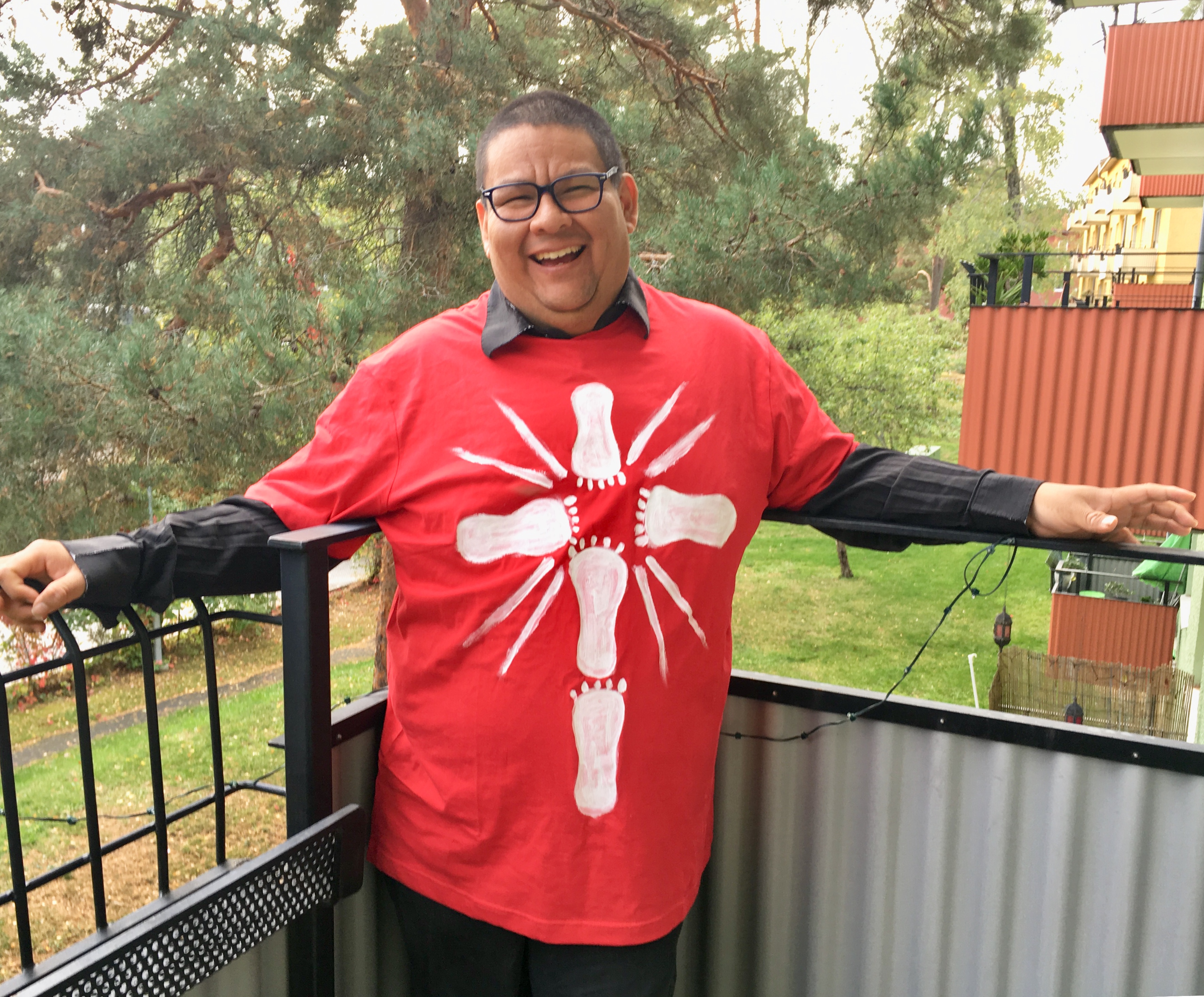 En person står och håller i ett räcke på en balkong. Hen ser glad ut. Det är grönt ute.