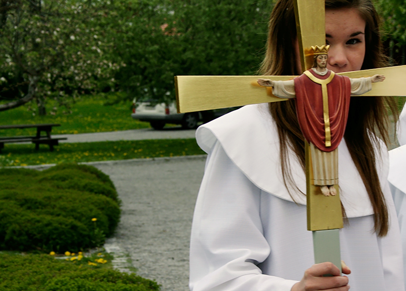 Konfirmand med processionskors med Kristusfigur på. Hennes ansikte är lite dolt bakom korset.